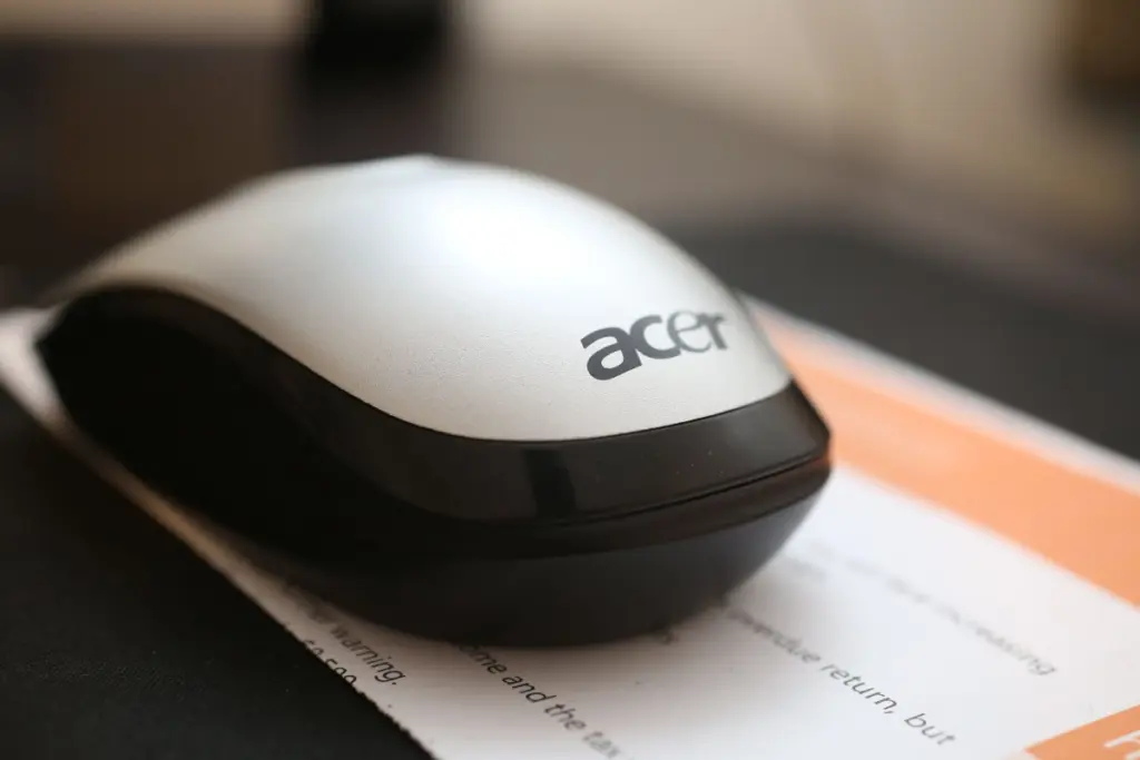 Close-up photograph of a silver and black Acer wireless computer mouse placed on a document. The Acer logo is